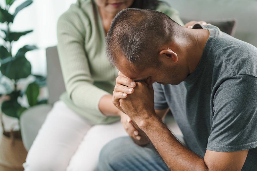 Belasteter Mann sitzt mit gesenktem Kopf auf dem Sofa, während eine Frau ihm tröstend die Hand hält.