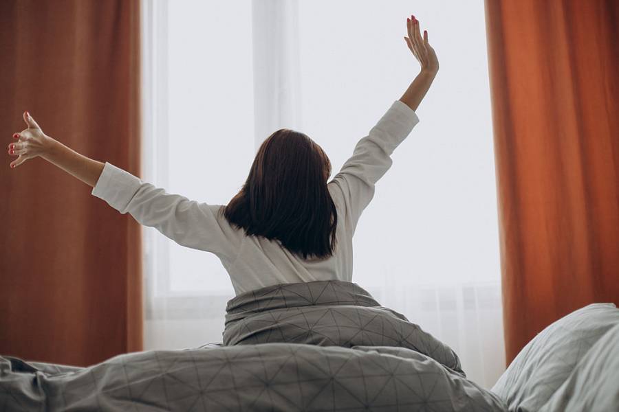 Frau streckt sich morgens im Bett vor hellem Fenster und startet entspannt in den Tag.